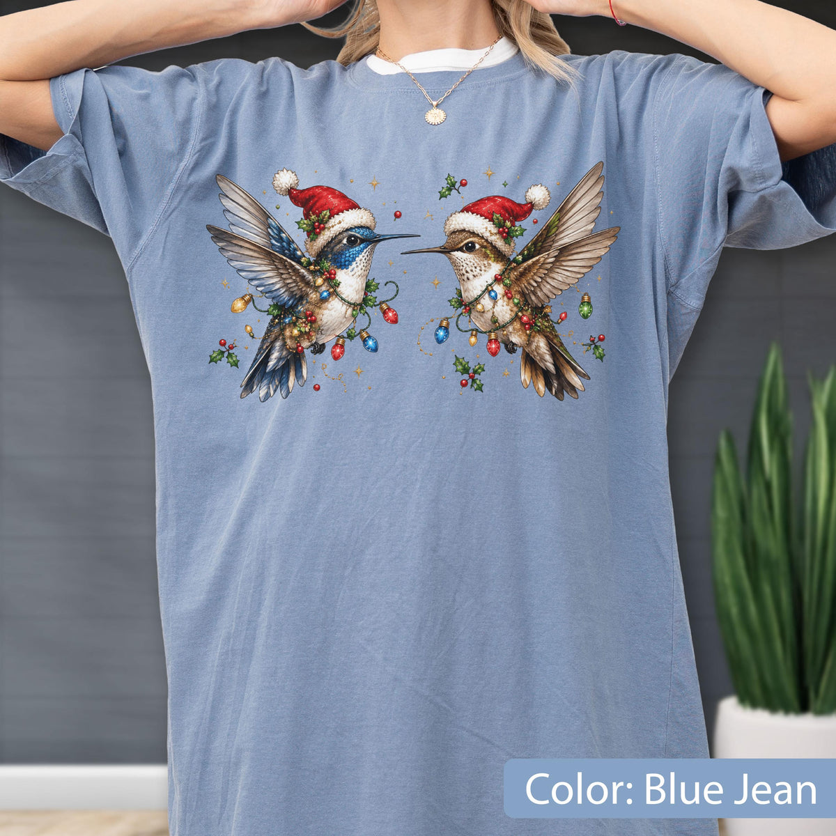 May include: A blue jean colored t-shirt featuring a graphic print of two hummingbirds wearing red and white Santa hats. The birds are surrounded by colorful Christmas light bulbs and green holly leaves, creating a festive holiday design on the front.