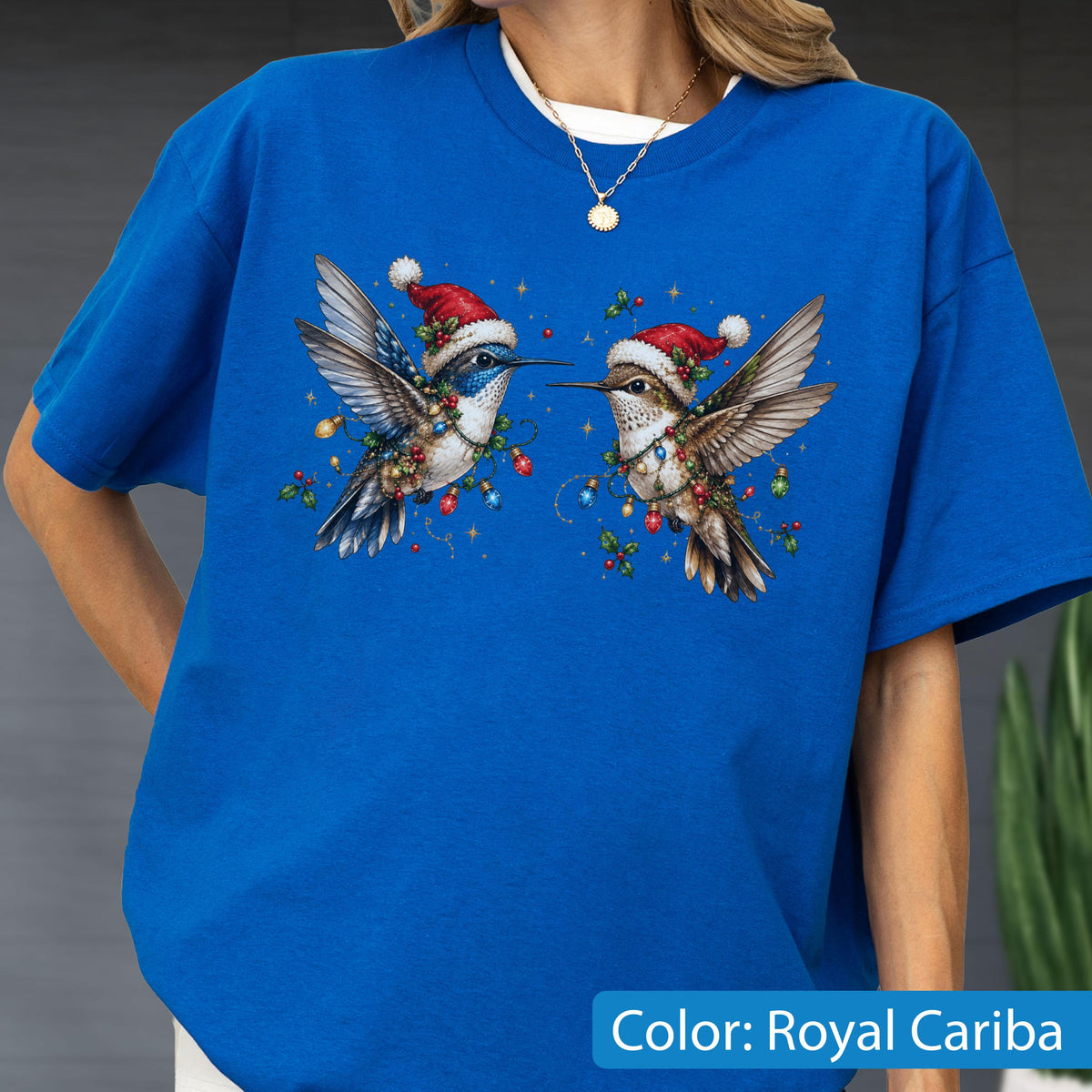 May include: A royal blue t-shirt featuring a graphic of two hummingbirds wearing red and white Santa hats. The birds are wrapped in colorful Christmas light strings and holly berries, with small gold stars scattered around the central design.