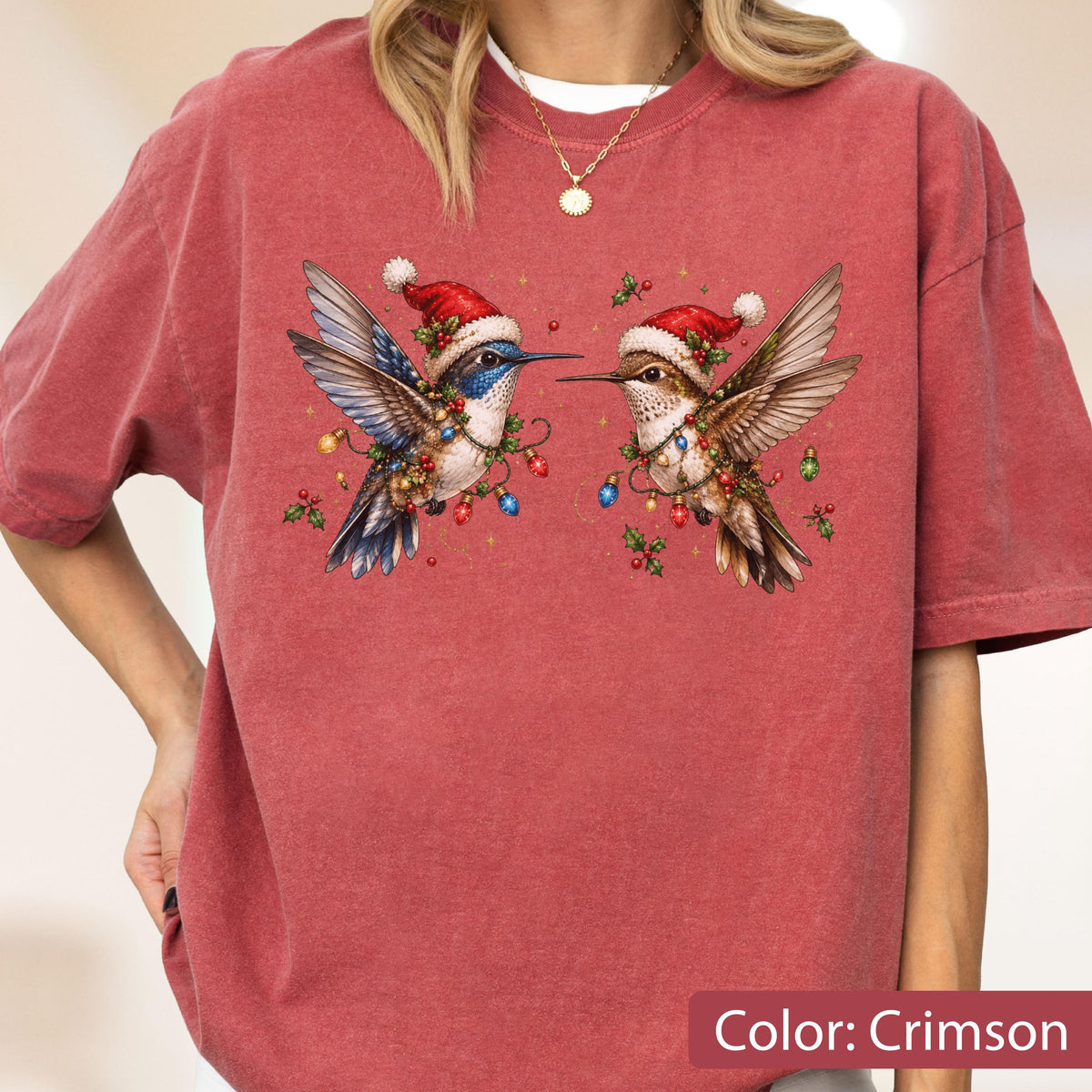 May include: A crimson short-sleeve t-shirt featuring a graphic of two hummingbirds wearing red Santa hats. The birds are decorated with colorful Christmas light strings and holly berries. The shirt has a crew neckline and a relaxed fit.