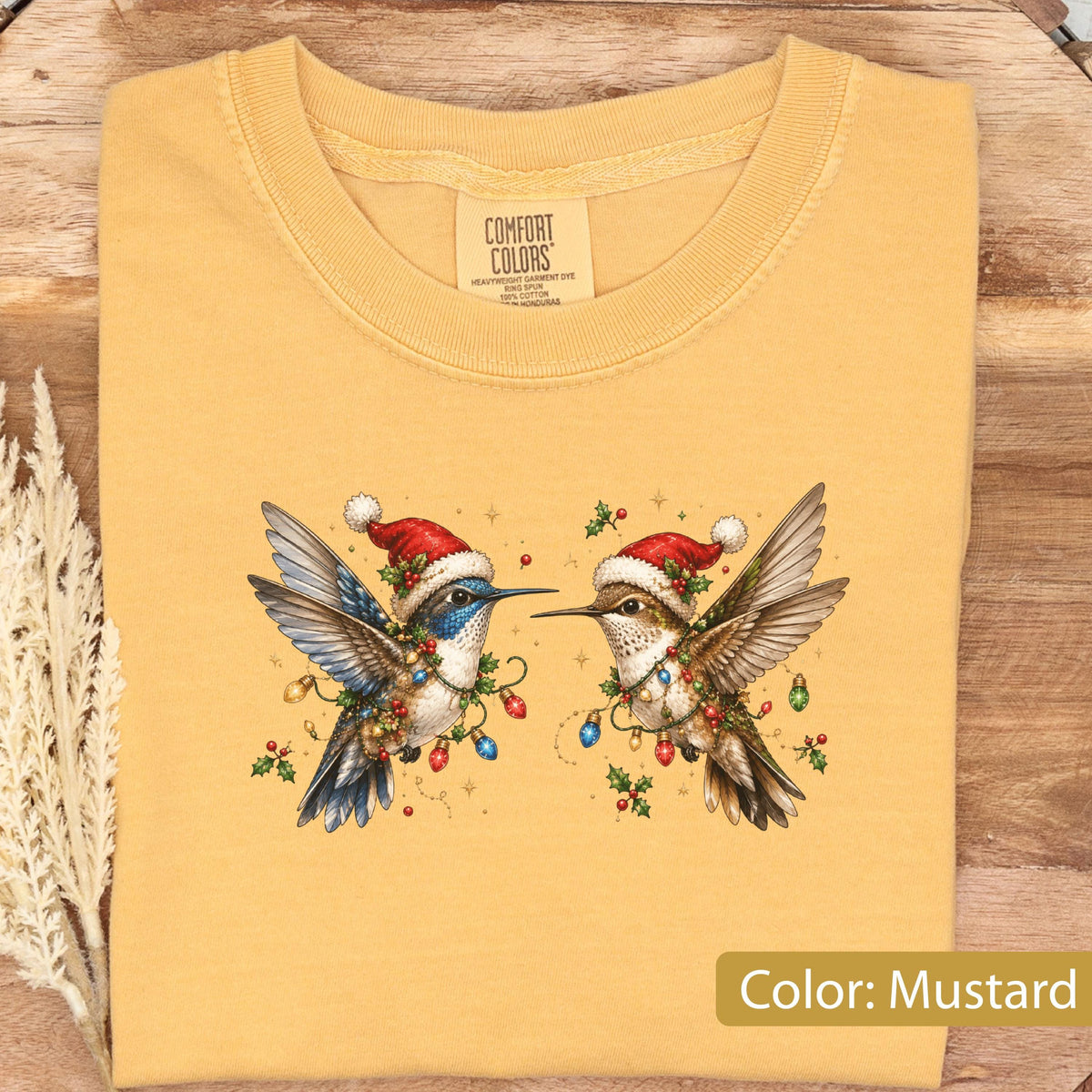 May include: A mustard yellow t-shirt featuring a graphic of two hummingbirds wearing red and white Santa hats. The birds are decorated with colorful Christmas light bulbs and holly berries. The inner neck label reads Comfort Colors.