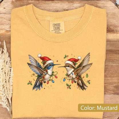 May include: A mustard yellow t-shirt featuring a graphic of two hummingbirds wearing red and white Santa hats. The birds are decorated with colorful Christmas light bulbs and holly berries. The inner neck label reads Comfort Colors.
