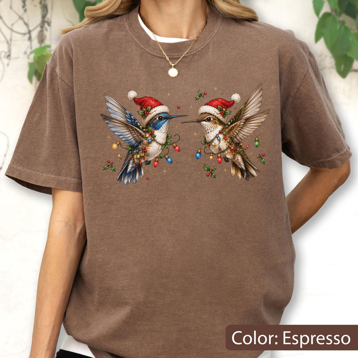 May include: An espresso brown t-shirt featuring a graphic of two hummingbirds wearing red and white Santa hats. The birds are decorated with colorful Christmas lights and green holly leaves. The shirt has a crew neckline and short sleeves.