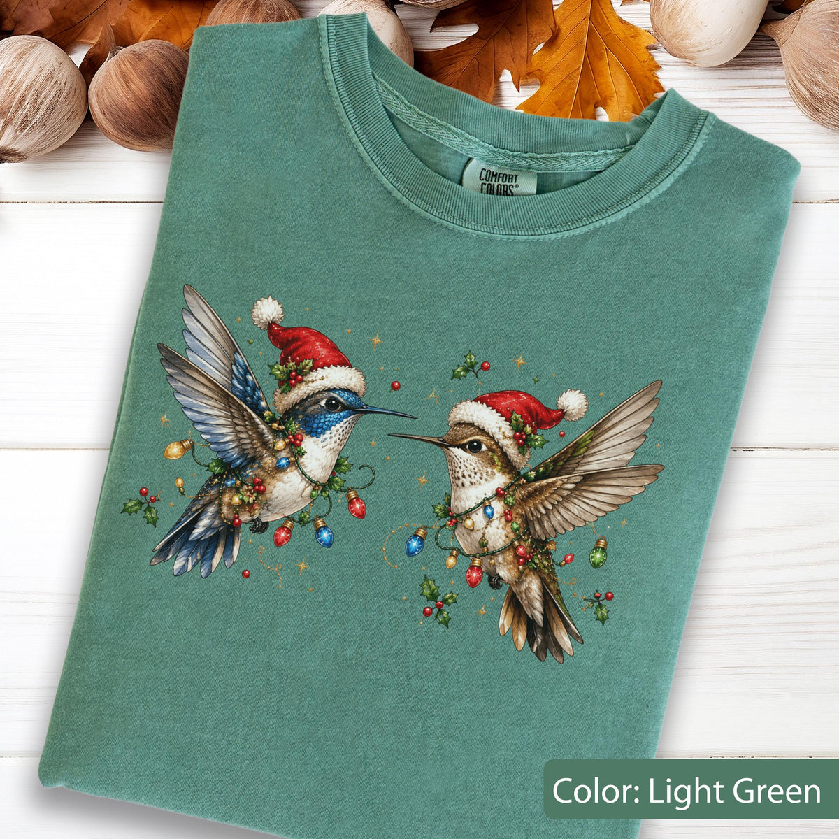 May include: A light green crew neck t-shirt featuring a graphic of two hummingbirds wearing red and white Santa hats. The birds are surrounded by colorful Christmas light bulbs and holly leaves. The shirt has a Comfort Colors brand label.