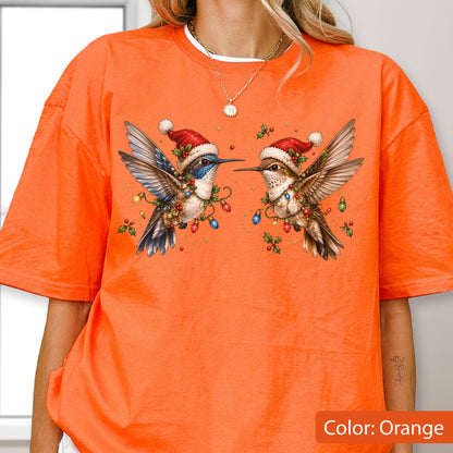 May include: An orange t-shirt featuring a graphic of two hummingbirds facing each other. Both birds wear red Santa hats with white pom-poms and are adorned with colorful Christmas light strings and small green holly leaves with red berries.