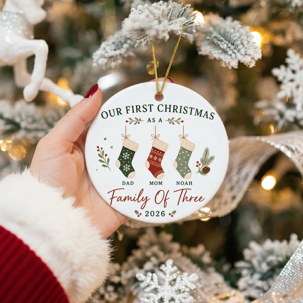 May include: A round white ceramic ornament featuring the text OUR FIRST CHRISTMAS AS A FAMILY OF THREE 2026. The design includes three hanging stockings labeled DAD, MOM, and NOAH, decorated with green, red, and sage patterns and pine sprigs.
