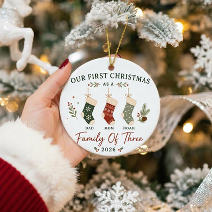 May include: A round white ceramic ornament featuring the text OUR FIRST CHRISTMAS AS A FAMILY OF THREE 2026. The design includes three hanging stockings labeled DAD, MOM, and NOAH, decorated with green, red, and sage patterns and pine sprigs.