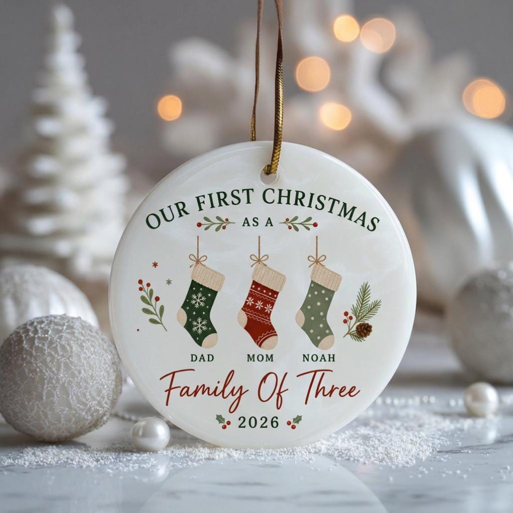 May include: A round white ceramic Christmas ornament featuring the text "OUR FIRST CHRISTMAS AS A FAMILY OF THREE" and "2026". It displays three hanging stockings labeled "DAD", "MOM", and "NOAH" with green, red, and light green patterns.
