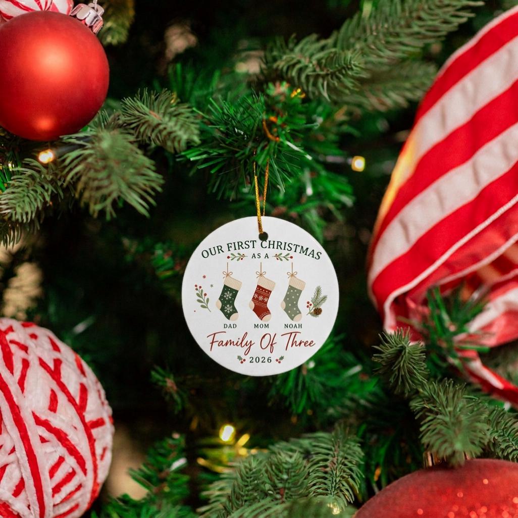 May include: A round white ceramic ornament featuring the text Our First Christmas As A Family Of Three 2026. The design includes three hanging stockings labeled Dad, Mom, and Noah, decorated with green and red patterns and small pine sprigs.