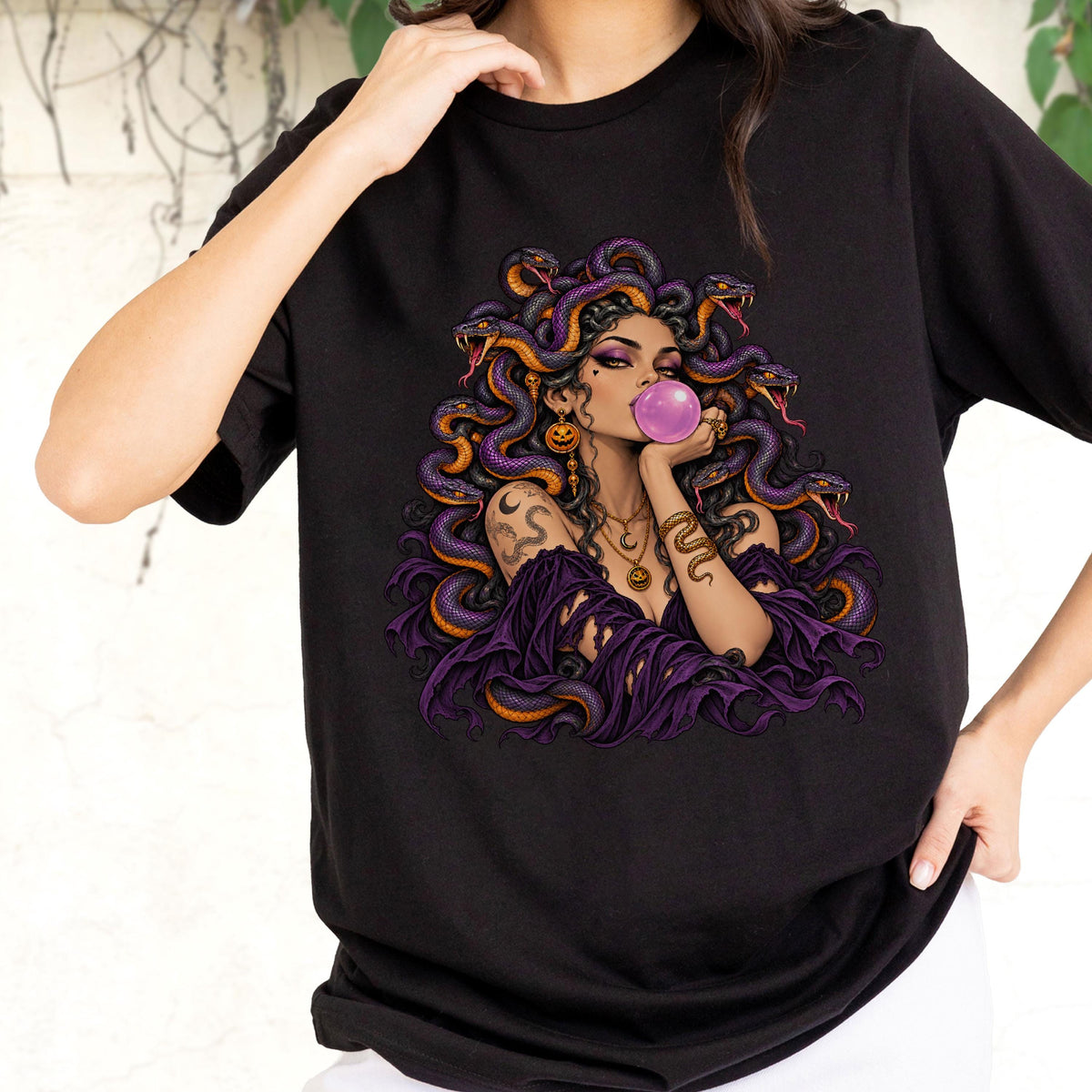 May include: A black t-shirt featuring a graphic illustration of a figure with snakes for hair, wearing a purple off-the-shoulder top, blowing a pink bubblegum bubble, and adorned with gold jewelry including a crescent moon and pumpkin charms.
