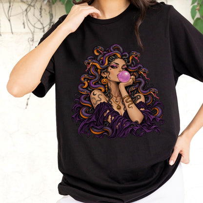 May include: A black t-shirt featuring a graphic illustration of a figure with snakes for hair, wearing a purple off-the-shoulder top, blowing a pink bubblegum bubble, and adorned with gold jewelry including a crescent moon and pumpkin charms.