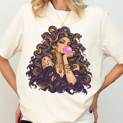 May include: A cream-colored t-shirt featuring a graphic illustration of a figure with purple snakes for hair, blowing a pink bubblegum bubble. The figure wears a dark purple off-the-shoulder garment, gold jewelry, and has a snake tattoo on the arm.