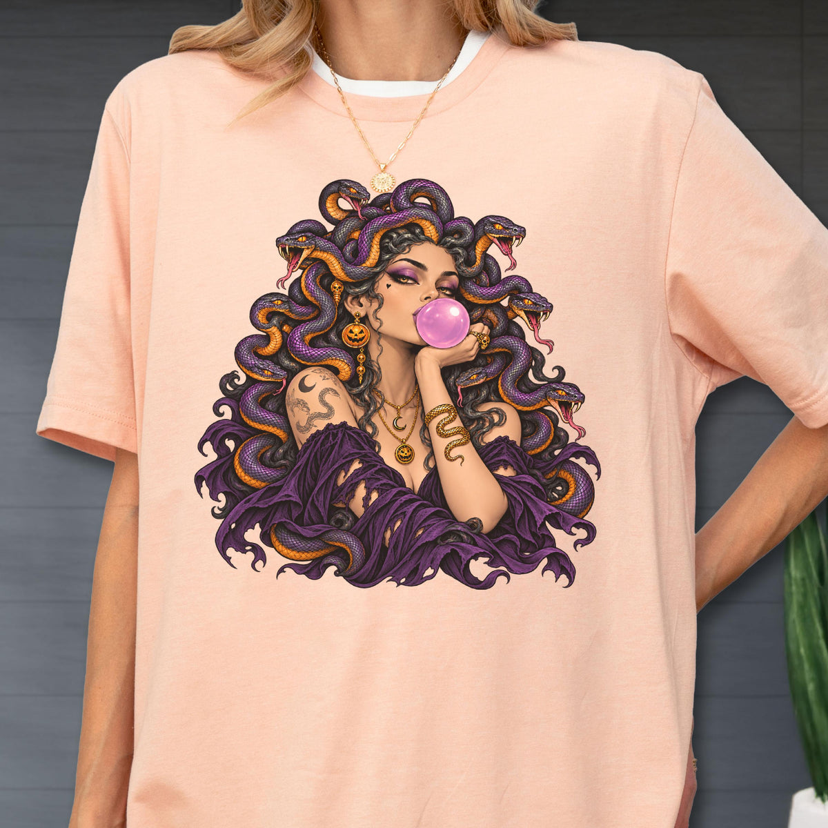 May include: A peach-colored t-shirt featuring a graphic illustration of a figure with purple snakes for hair, blowing a pink bubblegum bubble. The figure wears a dark purple garment, gold jewelry, and has a snake tattoo on the upper arm.