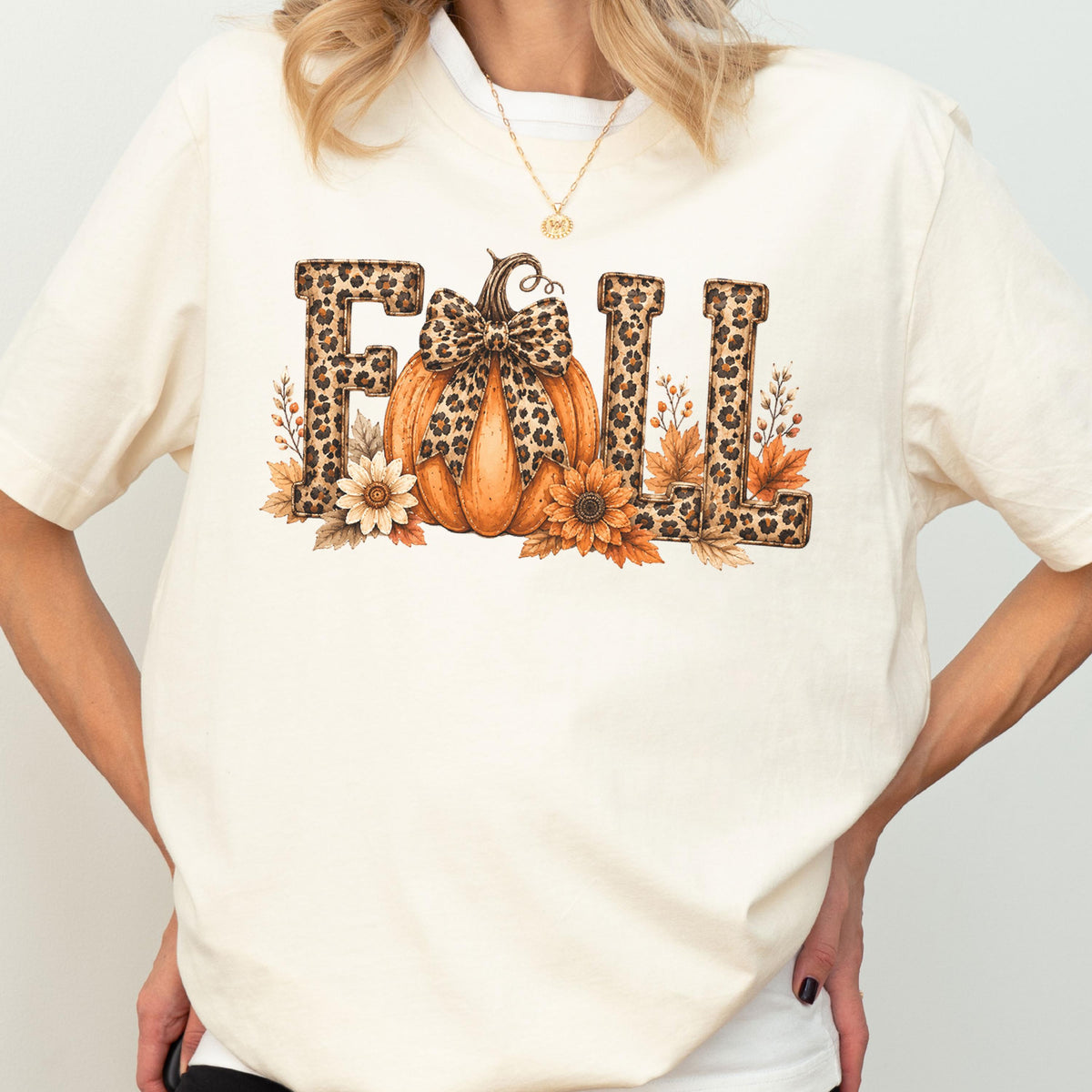 May include: A cream-colored t-shirt featuring a graphic print of the word FALL. The letters are filled with a brown leopard print pattern. An orange pumpkin with a leopard print bow sits in the center, surrounded by sunflowers and autumn leaves.