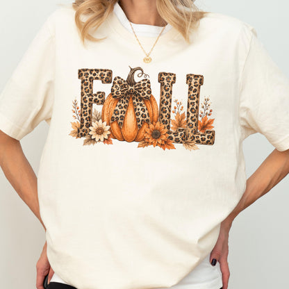 May include: A cream-colored t-shirt featuring a graphic print of the word FALL. The letters are filled with a brown leopard print pattern. An orange pumpkin with a leopard print bow sits in the center, surrounded by sunflowers and autumn leaves.