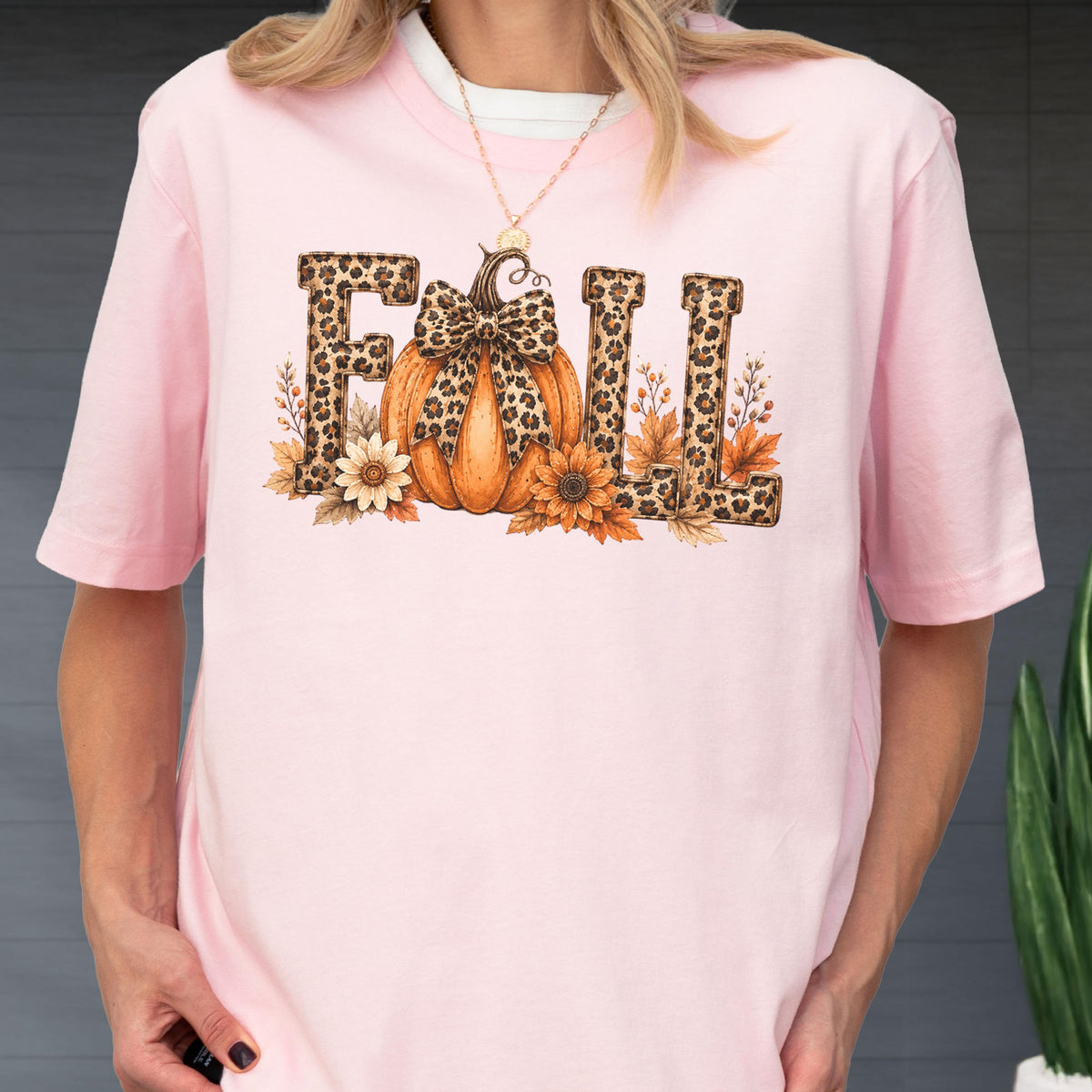 May include: A light pink short-sleeve t-shirt featuring a graphic print of the word FALL. The letters have a brown leopard print pattern. An orange pumpkin with a leopard print bow sits in the center, surrounded by brown and orange autumn leaves.