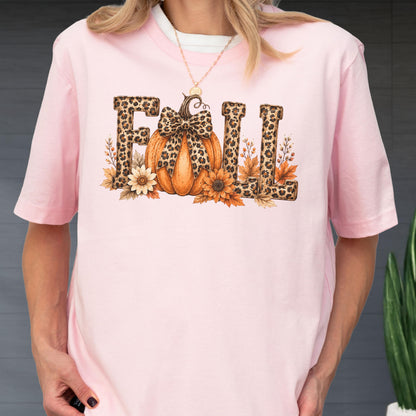 May include: A light pink short-sleeve t-shirt featuring a graphic print of the word FALL. The letters have a brown leopard print pattern. An orange pumpkin with a leopard print bow sits in the center, surrounded by brown and orange autumn leaves.