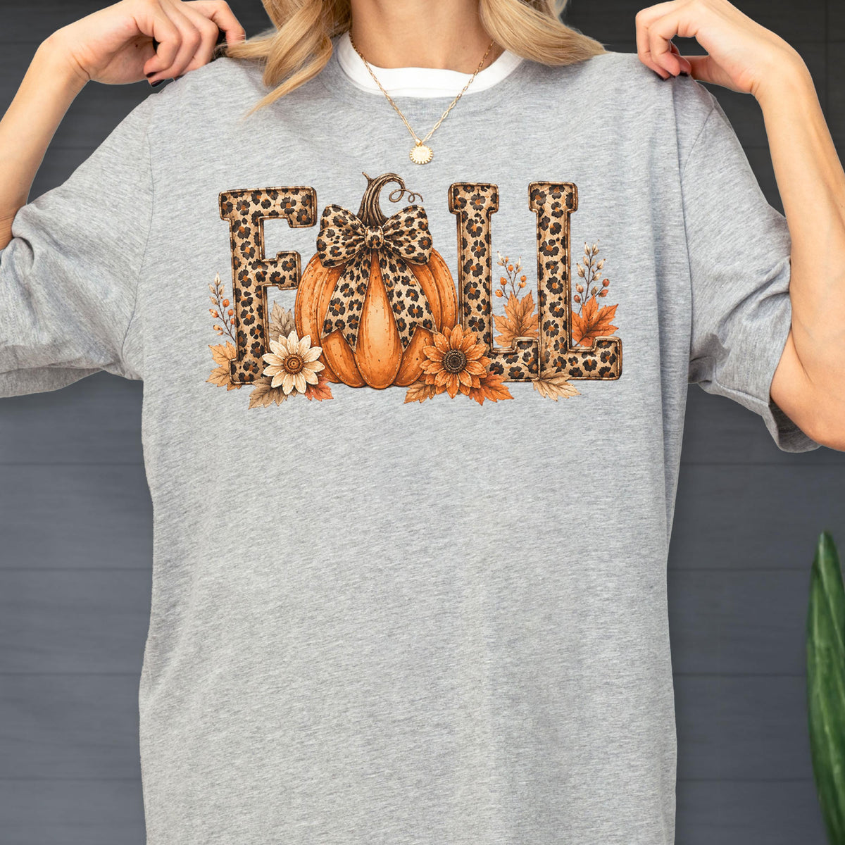 May include: A heather gray t-shirt featuring the word FALL in a leopard print pattern. The letter A is replaced by an orange pumpkin with a leopard print bow. The design includes sunflowers and autumn leaves surrounding the base of the letters.