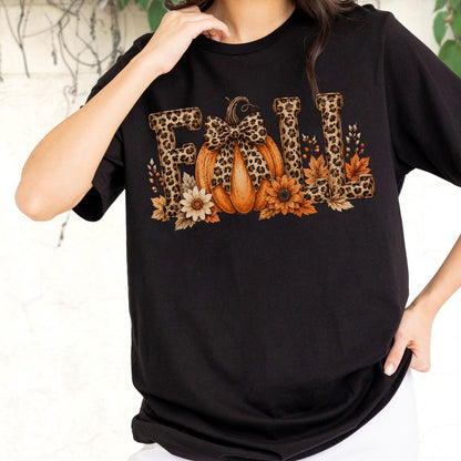 May include: A black short-sleeved t-shirt featuring a graphic print of the word FALL. The letters have a brown leopard print pattern. An orange pumpkin with a leopard print bow sits in the center, surrounded by brown and cream-colored flowers.