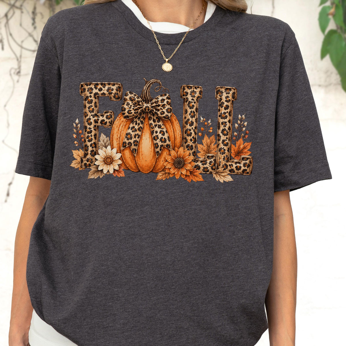 May include: A dark charcoal gray short-sleeve t-shirt featuring the word FALL in large block letters with a leopard print pattern. An orange pumpkin with a leopard print bow sits in the center, surrounded by orange and brown autumn leaves.