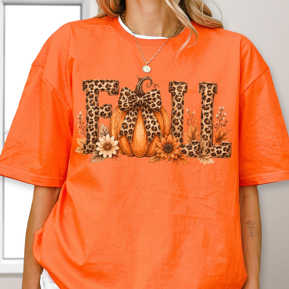 May include: A bright orange short-sleeve t-shirt featuring a graphic print of the word FALL. The letters have a leopard print pattern. A central orange pumpkin with a leopard print bow sits between the letters, surrounded by sunflowers.