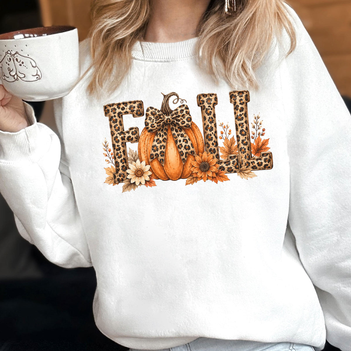 May include: A white crewneck sweatshirt featuring a graphic print of the word FALL in leopard print lettering. The letter A is replaced by an orange pumpkin with a leopard print bow, surrounded by sunflowers and autumn leaves in warm tones.