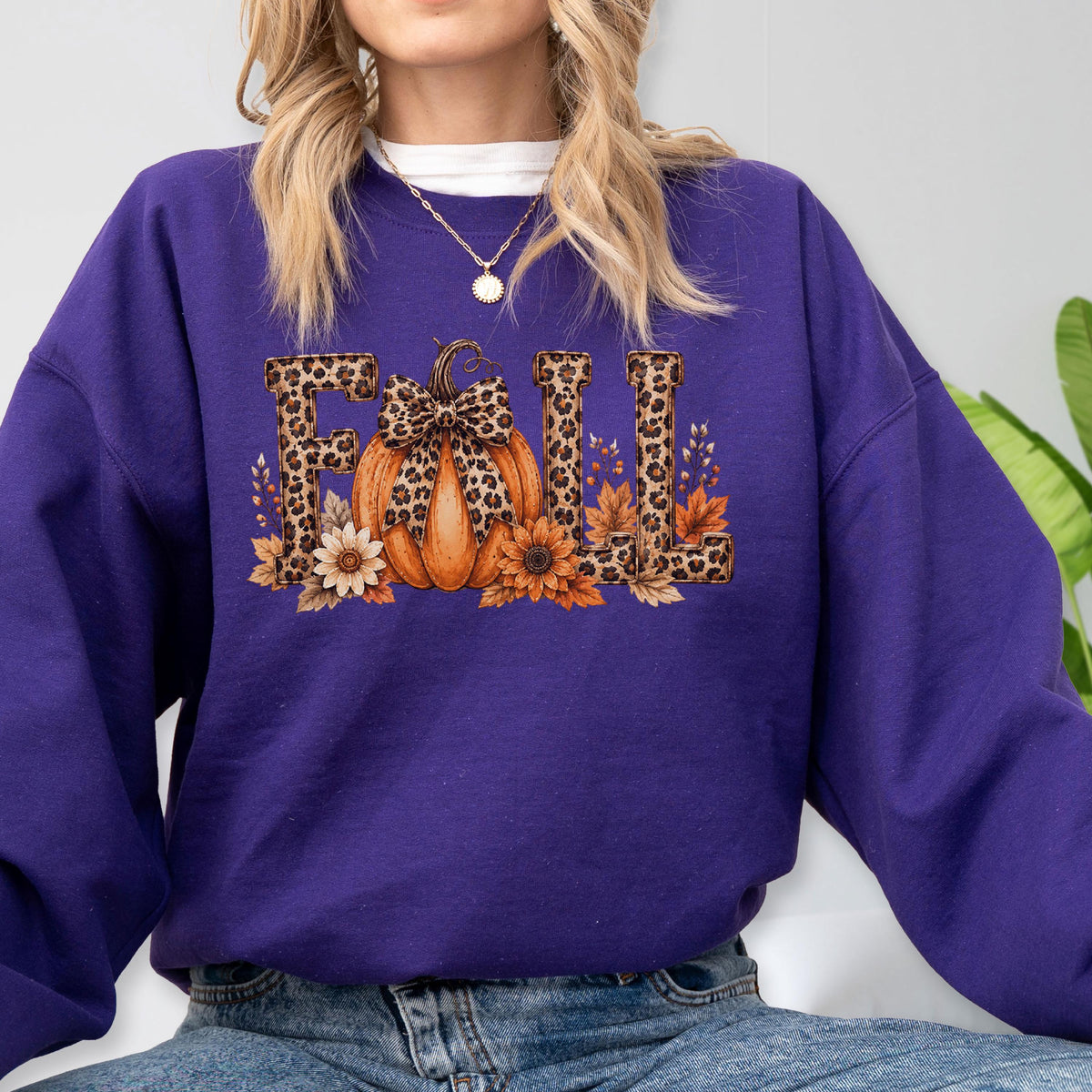 May include: A deep purple long-sleeve sweatshirt featuring a graphic print of the word FALL in leopard print letters. The letter A is replaced by an orange pumpkin with a leopard print bow, surrounded by sunflowers and autumn leaves.