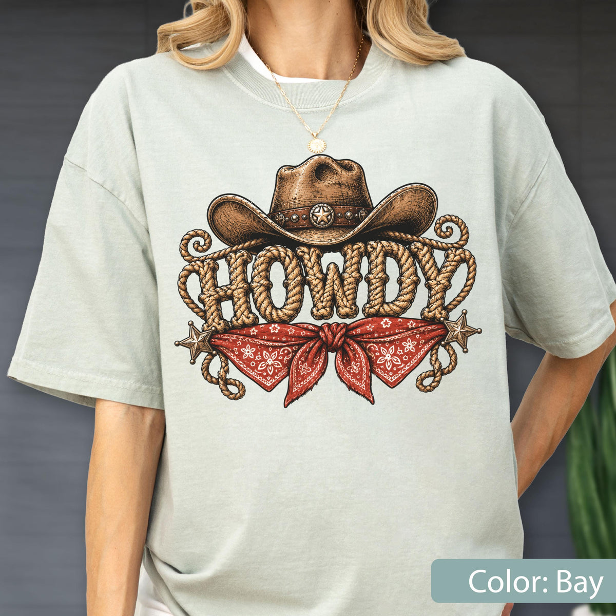 May include: A light sage green t-shirt featuring a graphic print of a brown cowboy hat above the word HOWDY written in a rope-style font. Below the text is a red bandana with a white floral pattern and two small metallic star accents.