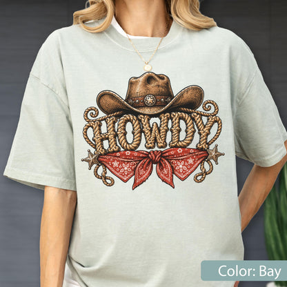 May include: A light sage green t-shirt featuring a graphic print of a brown cowboy hat above the word HOWDY written in a rope-style font. Below the text is a red bandana with a white floral pattern and two small metallic star accents.