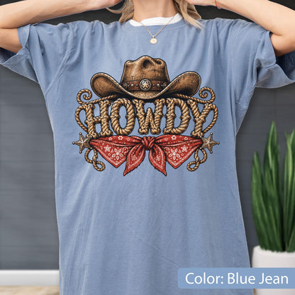 May include: A blue jean colored t-shirt featuring a graphic print of a brown cowboy hat, a red bandana with a white floral pattern, and the word HOWDY written in a rope-style font, accented by two small metallic stars on the left and right sides.