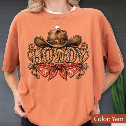 May include: A yam-colored t-shirt featuring a graphic print of a brown cowboy hat above the word HOWDY written in a rope-style font. Below the text is a red bandana with a white floral pattern, flanked by two small gold-colored stars.