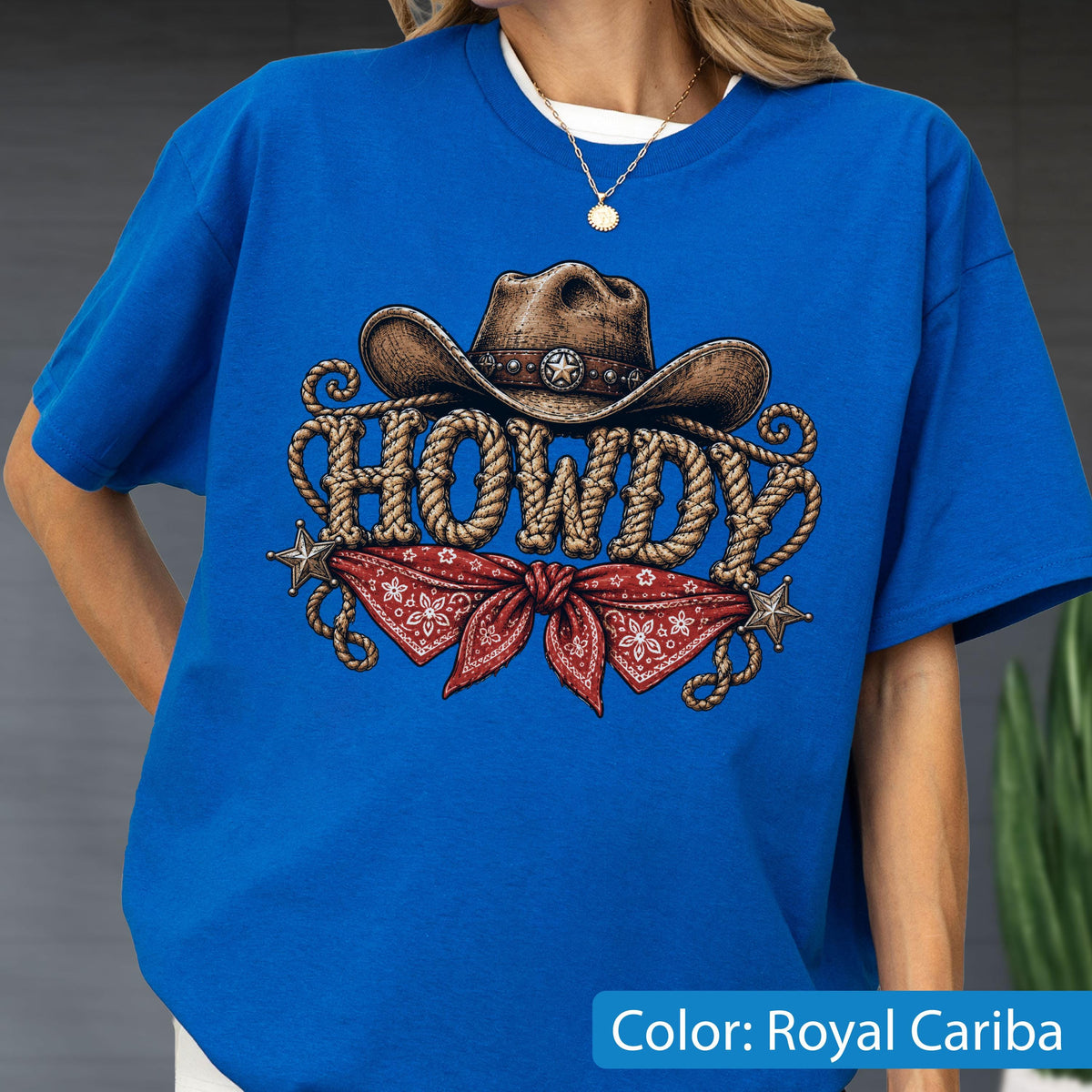 May include: A royal blue t-shirt featuring a graphic print of a brown cowboy hat above the word HOWDY written in a rope-style font. Below the text is a red bandana with a white floral pattern, flanked by two small silver-colored stars.