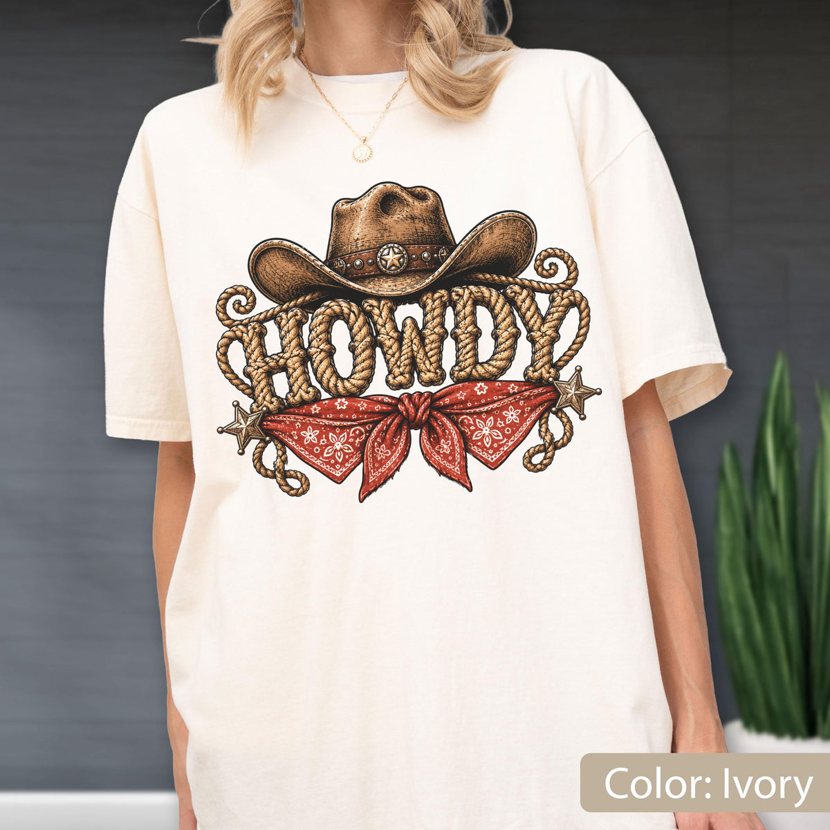 May include: An ivory t-shirt featuring a graphic print of a brown cowboy hat above the word HOWDY written in a rope-style font. Below the text is a red bandana with a white paisley pattern, flanked by two small brown stars on each side.