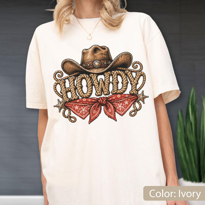 May include: An ivory t-shirt featuring a graphic print of a brown cowboy hat above the word HOWDY written in a rope-style font. Below the text is a red bandana with a white paisley pattern, flanked by two small brown stars on each side.