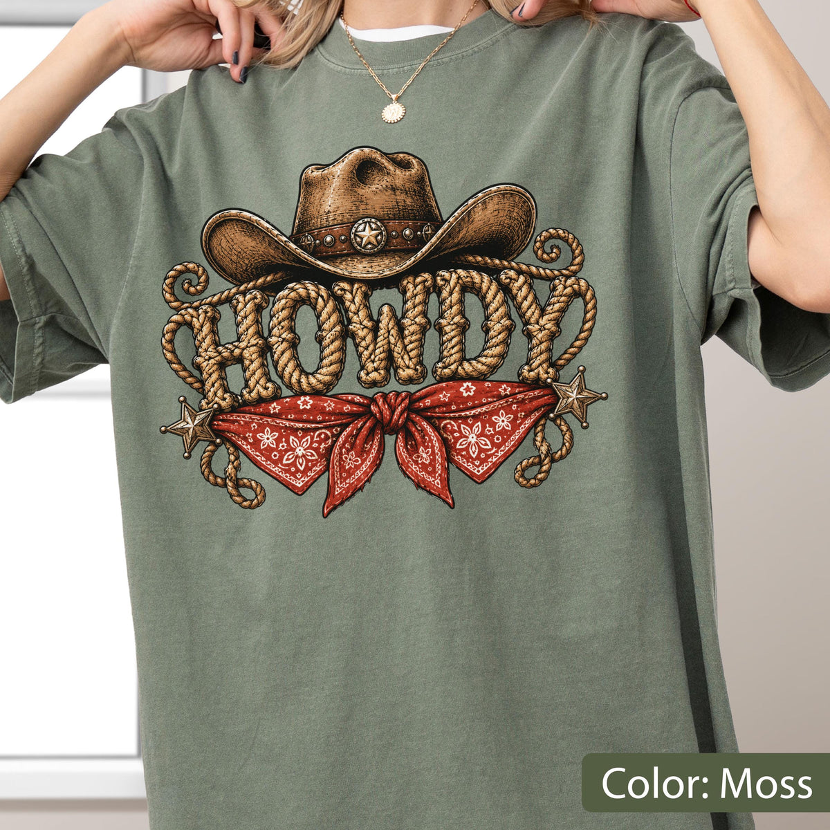 May include: A moss green t-shirt featuring a graphic print of a brown cowboy hat above the word HOWDY written in a rope-style font. Below the text is a red bandana with a white floral pattern, flanked by two small gold-colored stars.