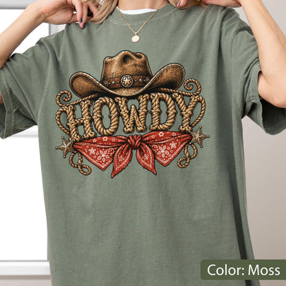 May include: A moss green t-shirt featuring a graphic print of a brown cowboy hat above the word HOWDY written in a rope-style font. Below the text is a red bandana with a white floral pattern, flanked by two small gold-colored stars.