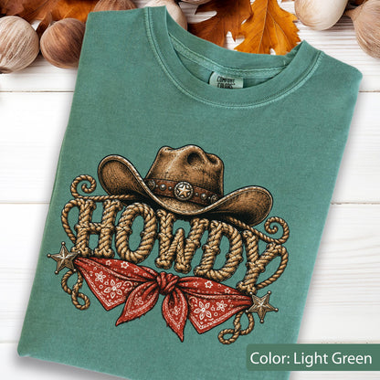 May include: A light green t-shirt featuring a graphic of a brown cowboy hat above the word HOWDY written in a rope-style font. Below the text is a red bandana with a white pattern and two small stars on the sides. The shirt has a Comfort Colors label.