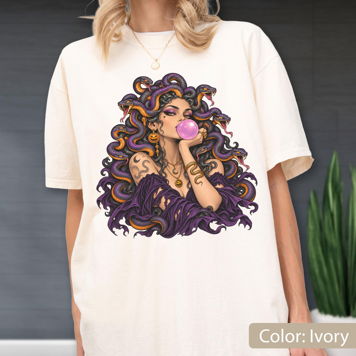 May include: An ivory t-shirt featuring a graphic illustration of a figure with purple snakes for hair, blowing a pink bubblegum bubble. The figure wears a dark purple off-the-shoulder garment, gold jewelry, and has a snake tattoo on the arm.