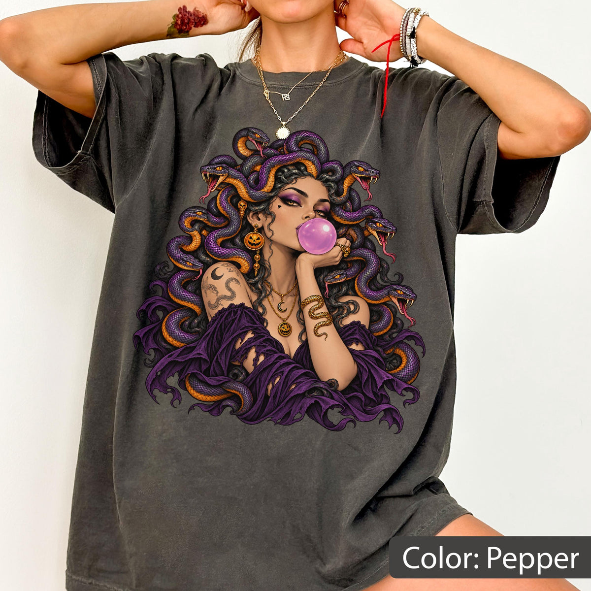 May include: A charcoal gray t-shirt featuring a graphic print of a figure with purple snakes for hair, blowing a pink bubblegum bubble. The figure wears a purple garment and gold jewelry, including a necklace and arm cuff, set against a dark background.