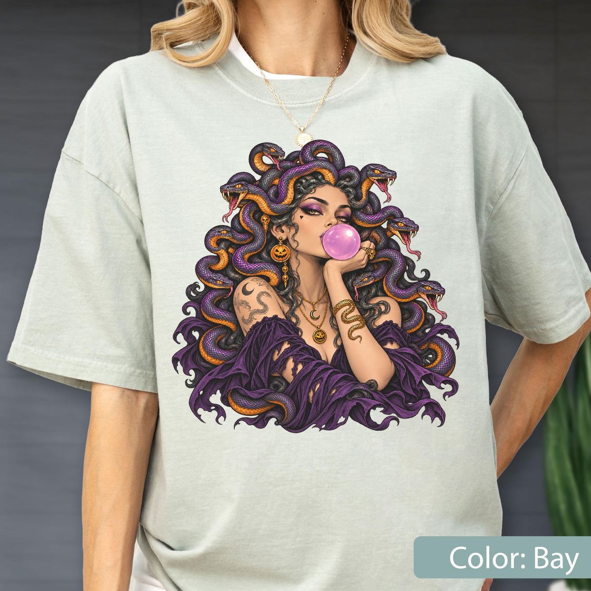 May include: A light sage green t-shirt featuring a graphic print of a figure with purple hair made of snakes, blowing a pink bubblegum bubble. The figure wears gold jewelry, including a necklace and earrings, and a dark purple off-shoulder garment.