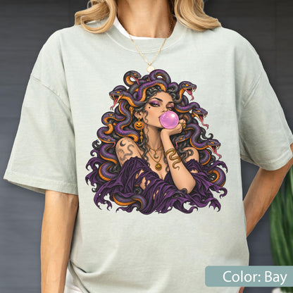 May include: A light sage green t-shirt featuring a graphic print of a figure with purple hair made of snakes, blowing a pink bubblegum bubble. The figure wears gold jewelry, including a necklace and earrings, and a dark purple off-shoulder garment.