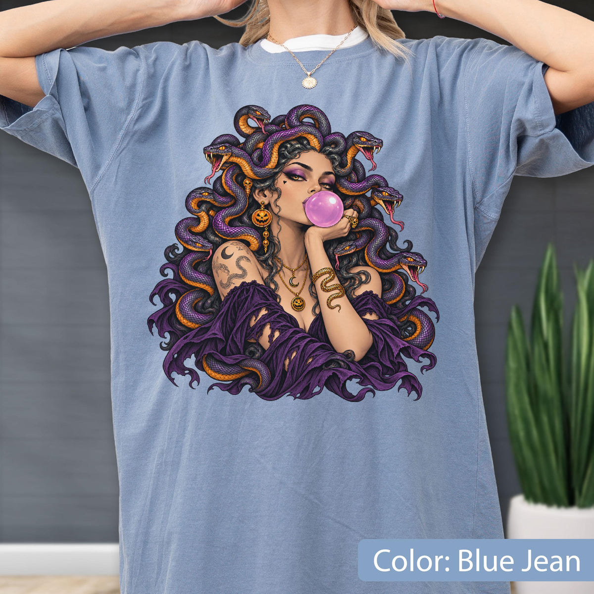 May include: A blue jean colored t-shirt featuring a graphic print of a figure with purple snakes for hair, blowing a pink bubble. The figure wears gold jewelry, including pumpkin-shaped earrings and a crescent moon necklace, with a purple garment.