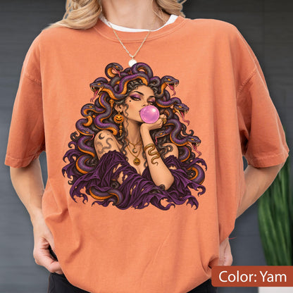 May include: An orange t-shirt featuring a graphic illustration of a figure with snakes for hair, wearing a purple garment, and blowing a pink bubble. The figure is adorned with gold-toned jewelry, including a necklace and earrings.