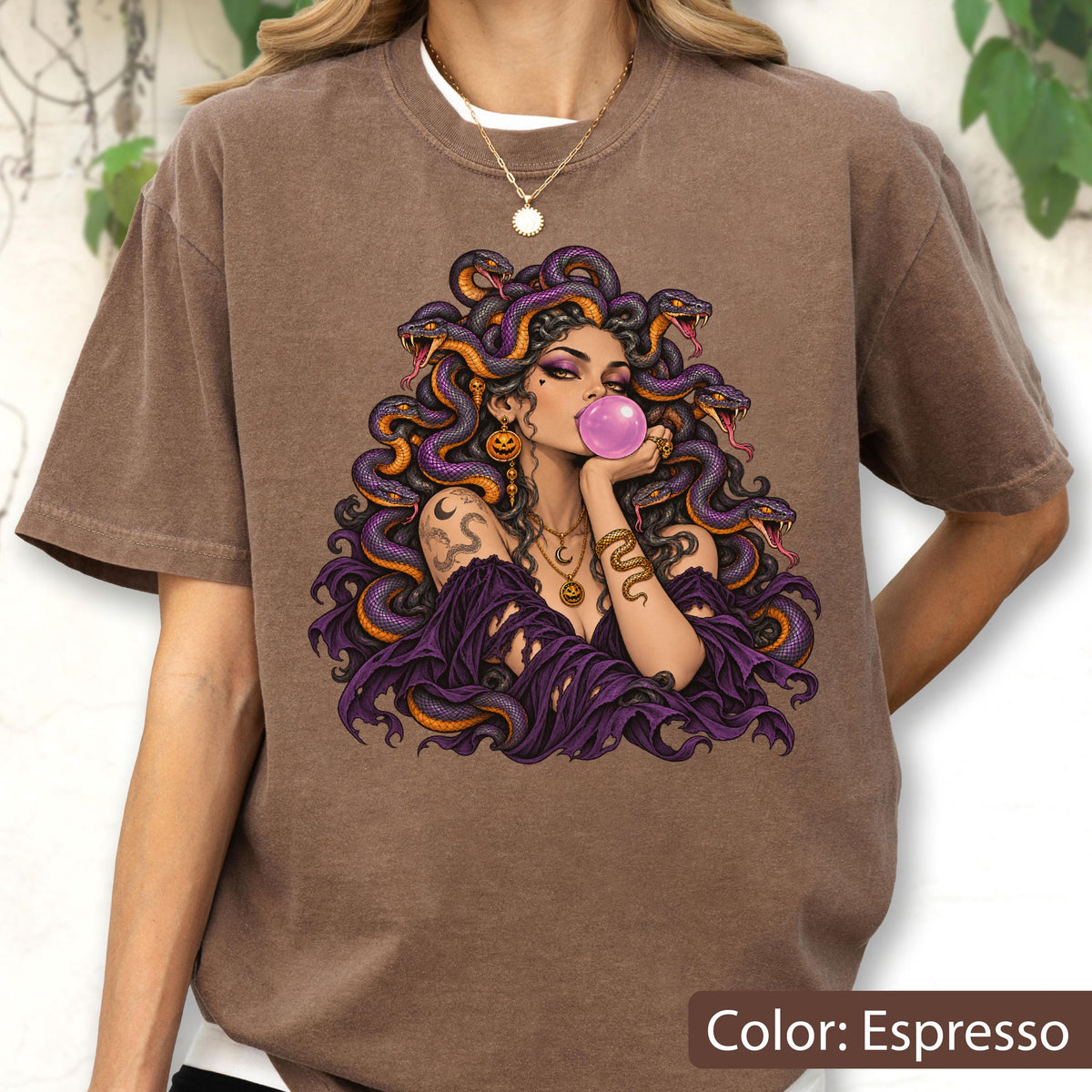May include: An espresso-colored t-shirt featuring a graphic illustration of a figure with purple snakes for hair, blowing a pink bubblegum bubble. The figure wears a dark purple off-the-shoulder garment, layered gold necklaces, and dangling pumpkin-shaped earrings.