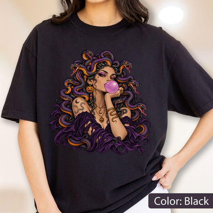 May include: A black t-shirt featuring a graphic illustration of a figure with purple snakes for hair, blowing a pink bubblegum bubble. The figure wears a purple off-the-shoulder top, layered gold necklaces, and has a snake tattoo on their arm.