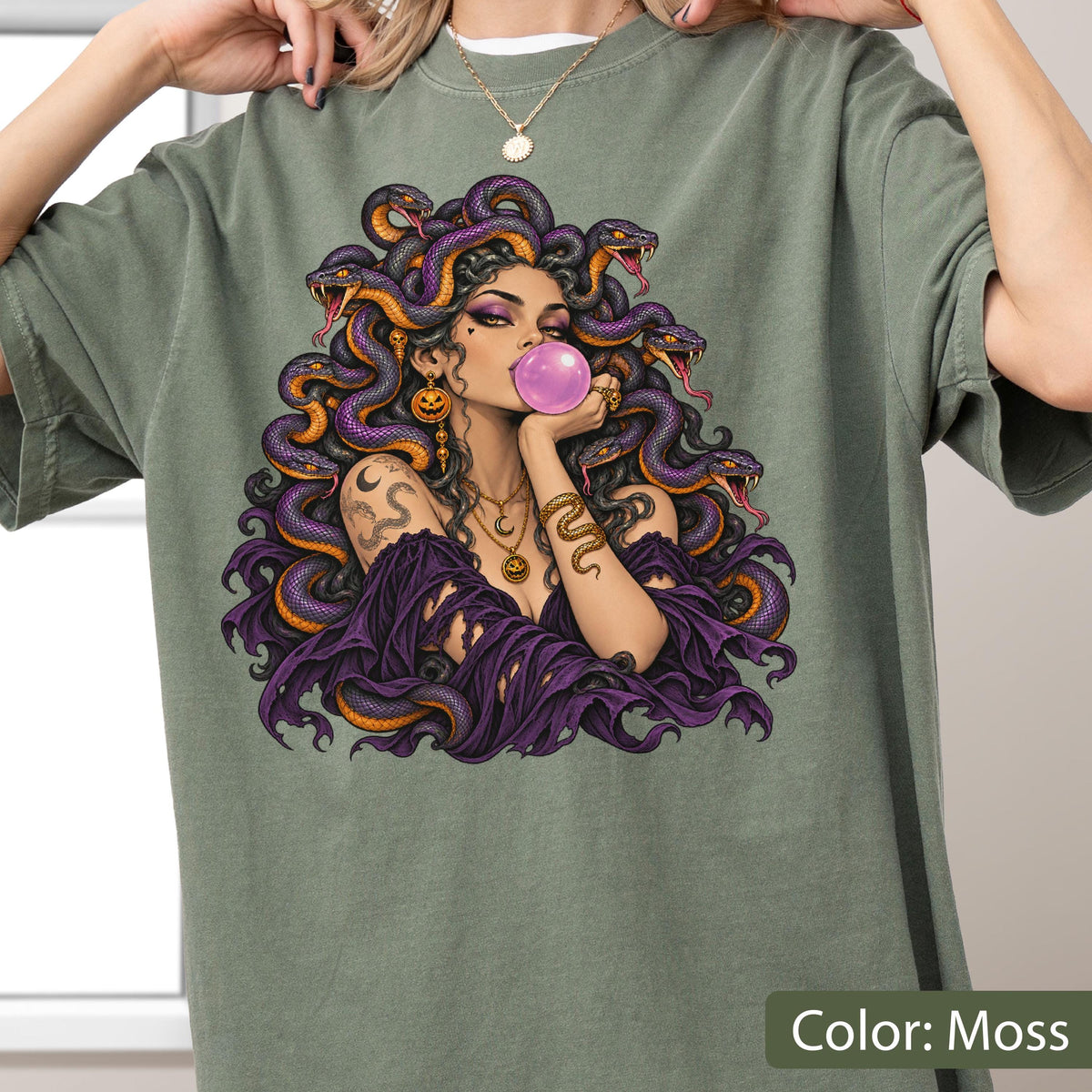 May include: A moss green t-shirt featuring a graphic illustration of a figure with purple snakes for hair, blowing a pink bubblegum bubble. The figure wears a dark purple off-the-shoulder garment, gold jewelry, and has a snake tattoo on the arm.