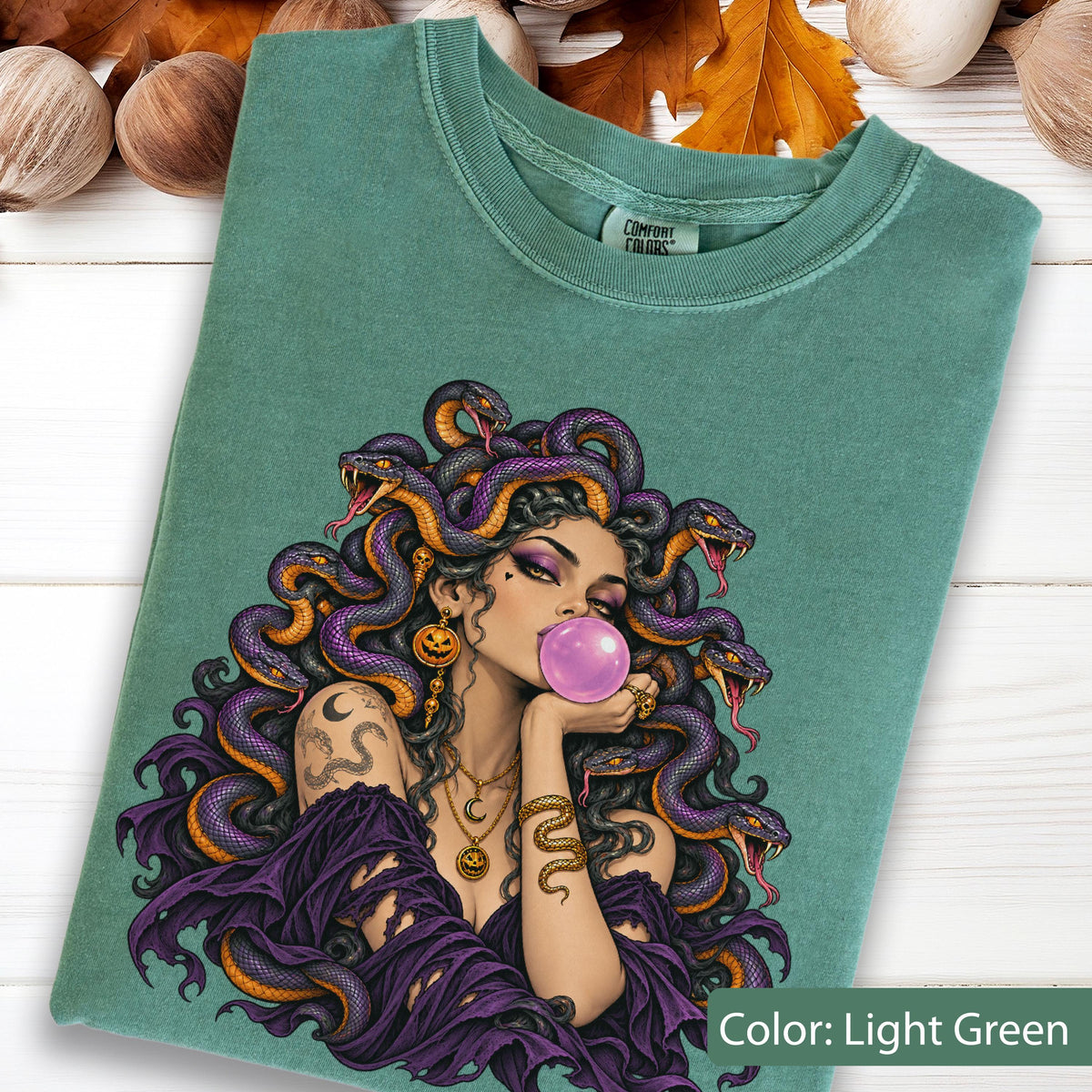 May include: A light green t-shirt featuring a graphic of a person with purple snakes for hair, blowing a pink bubblegum bubble. The person wears a purple garment, gold necklaces with pumpkin charms, and has a snake tattoo on their arm.