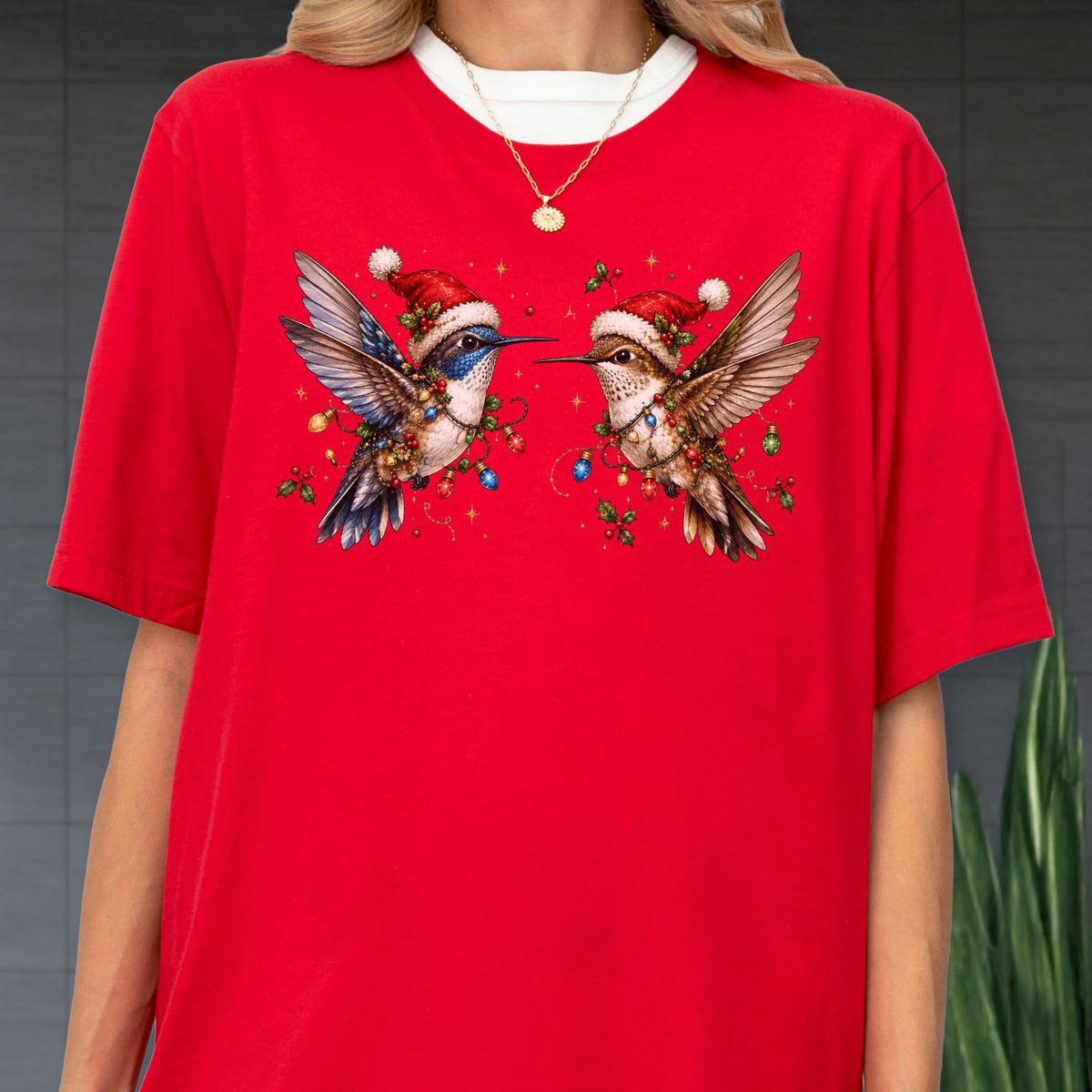 May include: A bright red t-shirt featuring a graphic of two hummingbirds facing each other. Each bird wears a small red Santa hat and is surrounded by colorful Christmas lights and green holly leaves. The design is centered on the chest area.