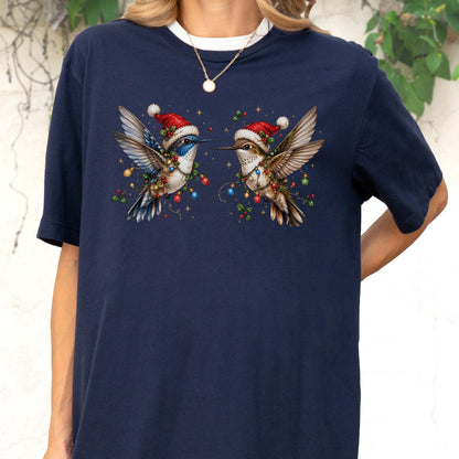 May include: A navy blue short-sleeve t-shirt featuring a graphic of two hummingbirds wearing red and white Santa hats. The birds are wrapped in colorful Christmas light strings and holly berries, with a white crew neck collar visible at the top.