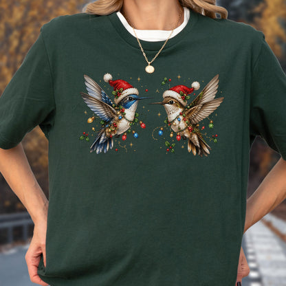 May include: A dark forest green t-shirt featuring a graphic of two hummingbirds wearing red Santa hats. The birds are adorned with colorful Christmas light strings and holly berries, set against a dark green fabric background with a crew neckline.