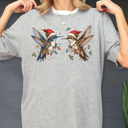 May include: A heather gray t-shirt featuring a graphic of two hummingbirds wearing red and white Santa hats. The birds are surrounded by colorful Christmas light bulbs, holly leaves, and red berries, creating a festive holiday-themed illustration.